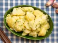 Un tazón verde lleno de patatas alioli cremosas, adornadas con perejil fresco. A su lado, ajos y tenedores de madera sobre un mantel de cuadros azules.