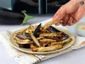 Berenjenas crujientes en freidora de aire, espolvoreadas con sésamo. Una mano toma una rodaja dorada.