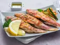 Cuatro salmonetes al horno, dorados y jugosos, servidos con rodajas de limón, perejil y patatas fritas.
