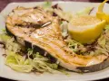 Pez espada a la plancha con ajo y perejil, servido sobre una cama de lechugas y acompañado de un gajo de limón.