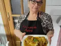 Una mujer sonriente muestra un plato de manitas de cerdo en salsa. Lleva un delantal negro con el texto "La Cocina de Adora".
