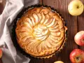 Tarta de manzana dorada, decorada con láminas de fruta, rodeada de manzanas frescas y espigas de trigo sobre una mesa de madera.