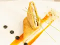 Un crujiente san jacobo, cortado por la mitad, reposa sobre una salsa naranja. Brotes y puntos oscuros decoran el plato.