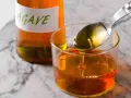 Una cuchara vierte sirope de agave dorado en un vaso lleno de este dulce líquido. Una botella con etiqueta "AGAVE" está al fondo.