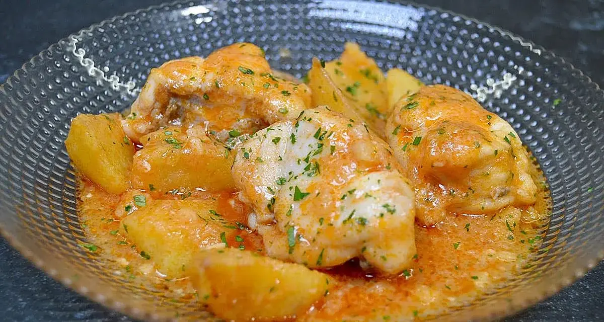 Rape a la marinera con patatas, servido en un cuenco de cristal con salsa naranja y perejil picado.
