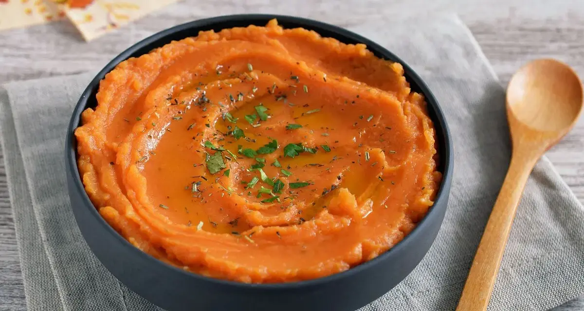 Un tazón de cremoso puré de boniato, adornado con aceite y hierbas, junto a una cuchara de madera.