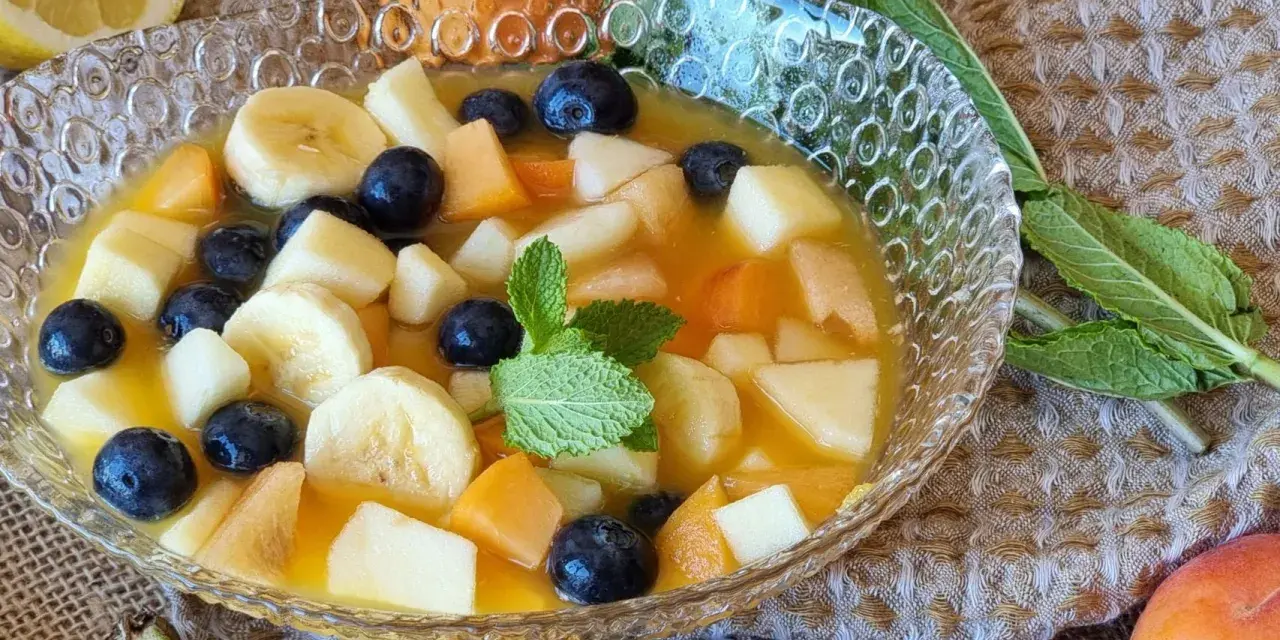 Deliciosa macedonia de frutas con plátano, arándanos y melocotón en un cuenco de cristal, decorada con menta.