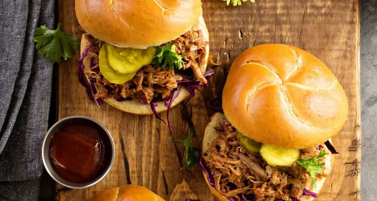 Dos sándwiches de pulled pork jugoso, con repollo morado, pepinillos y salsa barbacoa en un plato de madera.