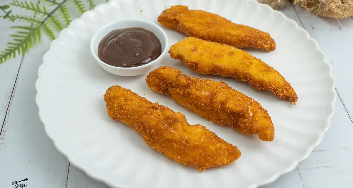 Cuatro crujientes fingers de pollo dorados, servidos en un plato blanco con una pequeña taza de salsa barbacoa.