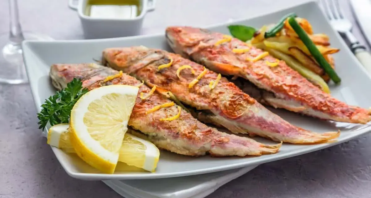 Cuatro salmonetes al horno, dorados y jugosos, servidos con rodajas de limón, perejil y patatas fritas.