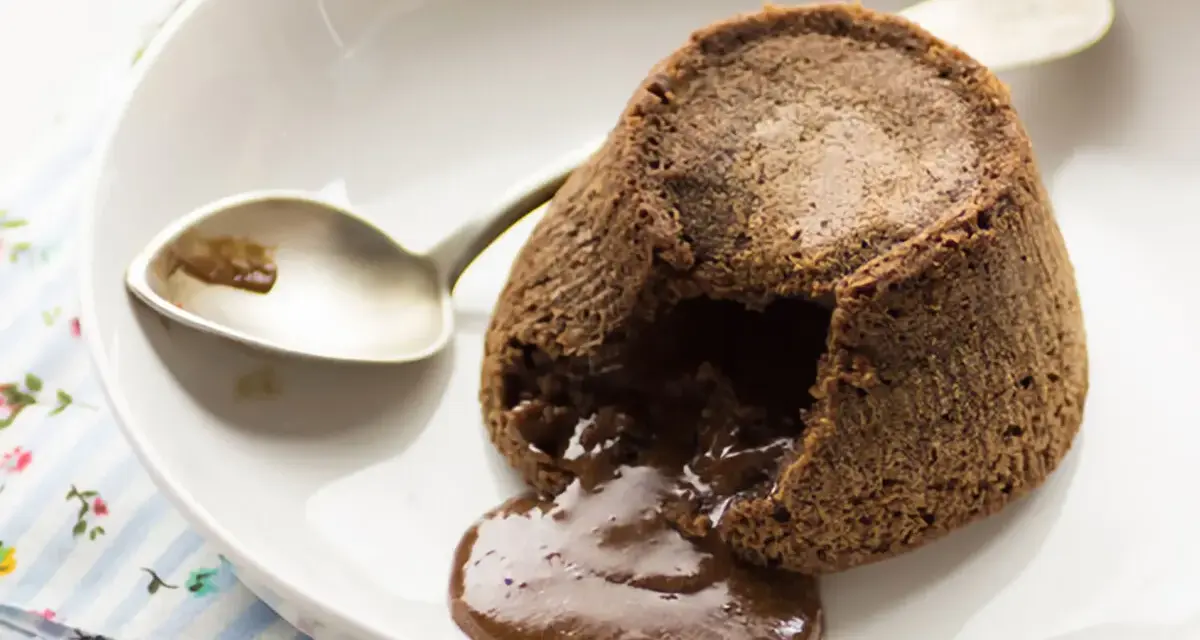 Coulant de chocolate recién hecho, con su interior líquido y cremoso desbordándose. Una cuchara plateada espera pacientemente.