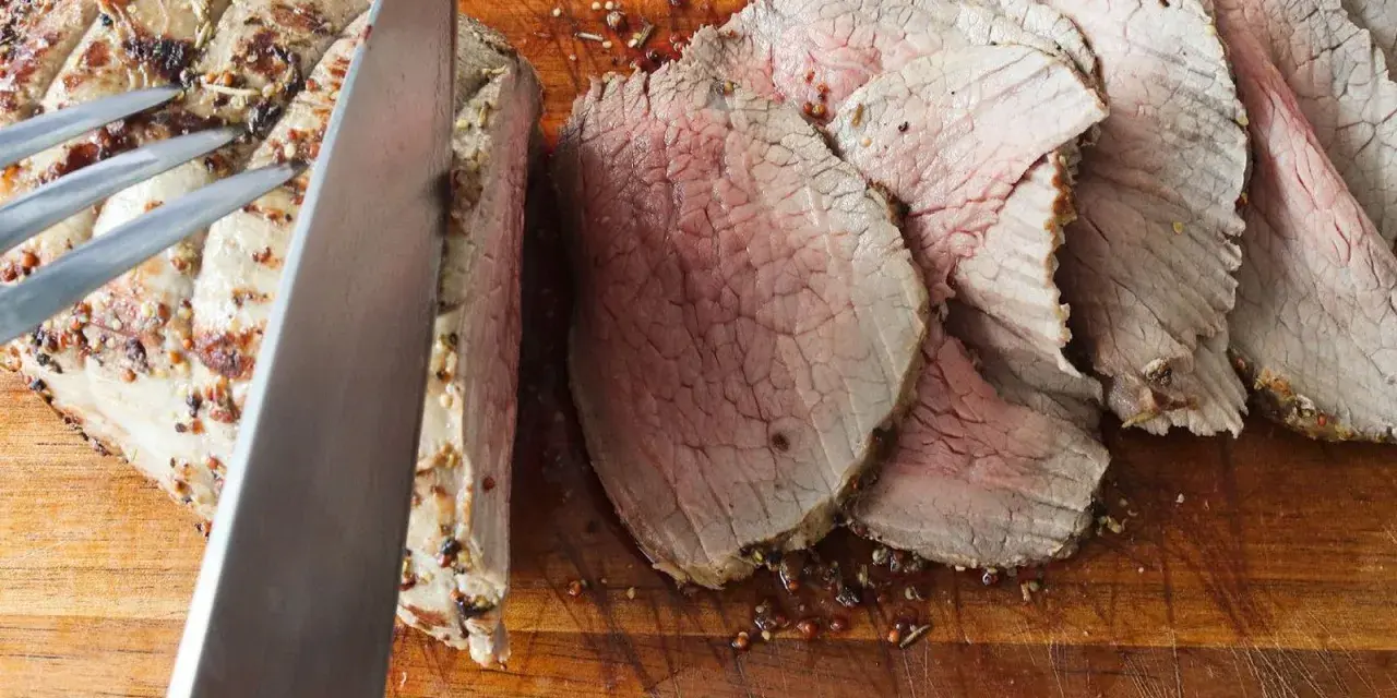 Un cuchillo corta un jugoso roast beef en rodajas sobre una tabla de madera.