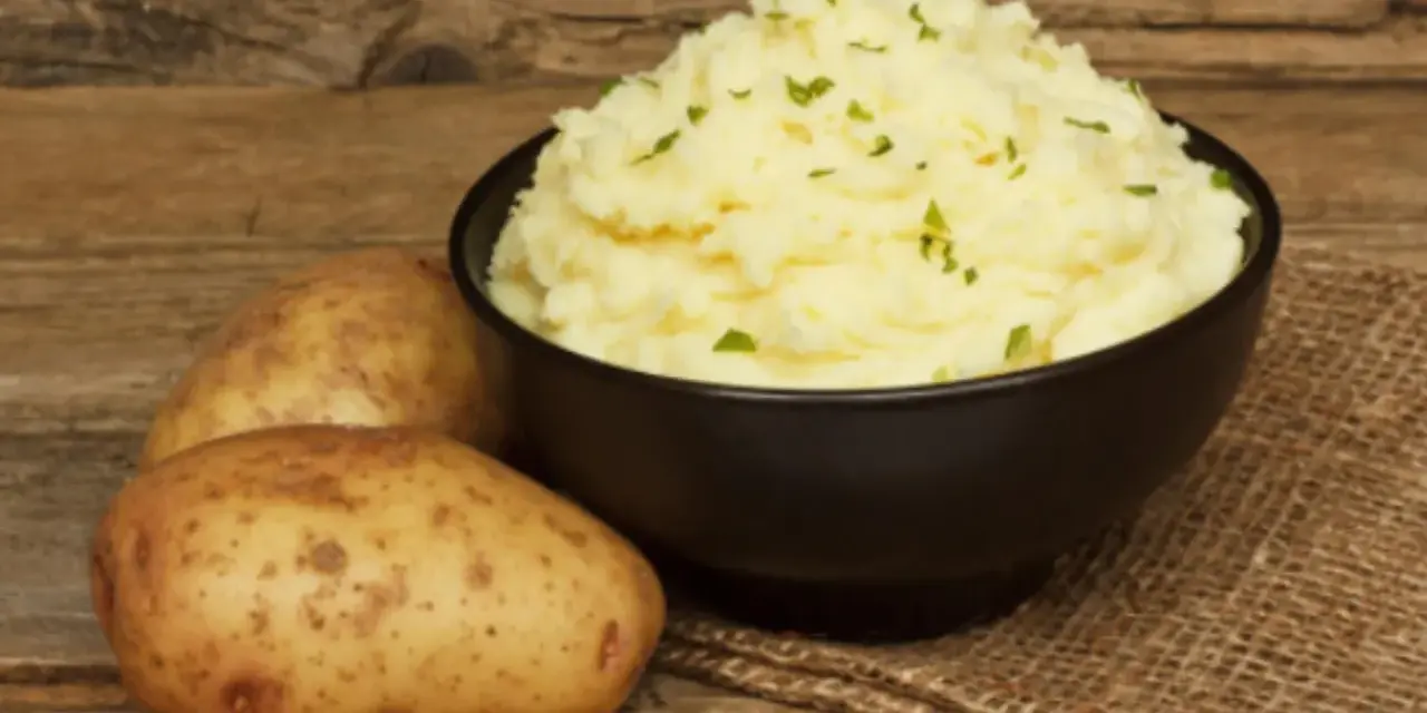 Un tazón de cremoso puré de patatas, adornado con perejil picado, junto a patatas enteras sobre una mesa de madera rústica.