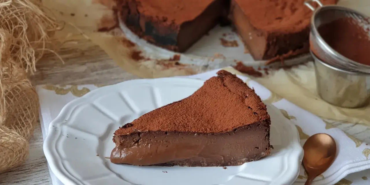 Una porción de tarta de chocolate, cubierta de cacao en polvo, reposa en un plato blanco. El resto de la tarta, también espolvoreada, se ve al fondo.