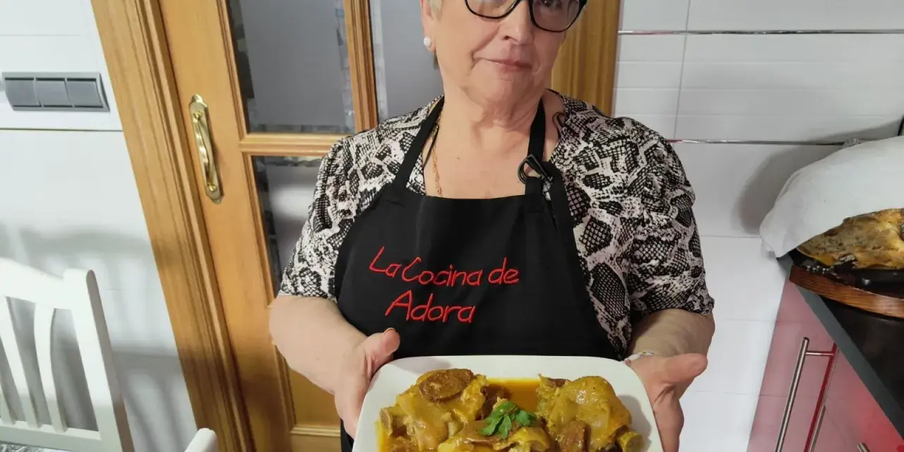 Una mujer sonriente muestra un plato de manitas de cerdo en salsa. Lleva un delantal negro con el texto "La Cocina de Adora".