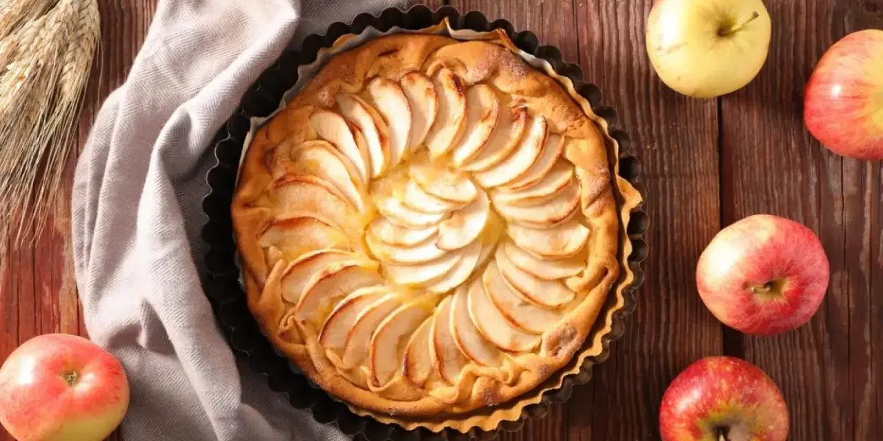 Tarta de manzana dorada, decorada con láminas de fruta, rodeada de manzanas frescas y espigas de trigo sobre una mesa de madera.