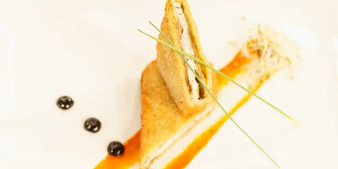 Un crujiente san jacobo, cortado por la mitad, reposa sobre una salsa naranja. Brotes y puntos oscuros decoran el plato.