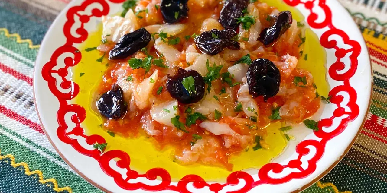 Esqueixada de bacalao con aceitunas negras y perejil, servida en un plato blanco con borde rojo sobre un mantel colorido.