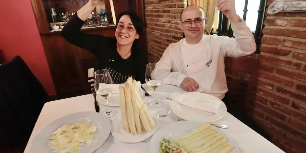 Pareja sonriente celebra con espárragos blancos en un restaurante. Platos de espárragos blancos y ensalada adornan la mesa.