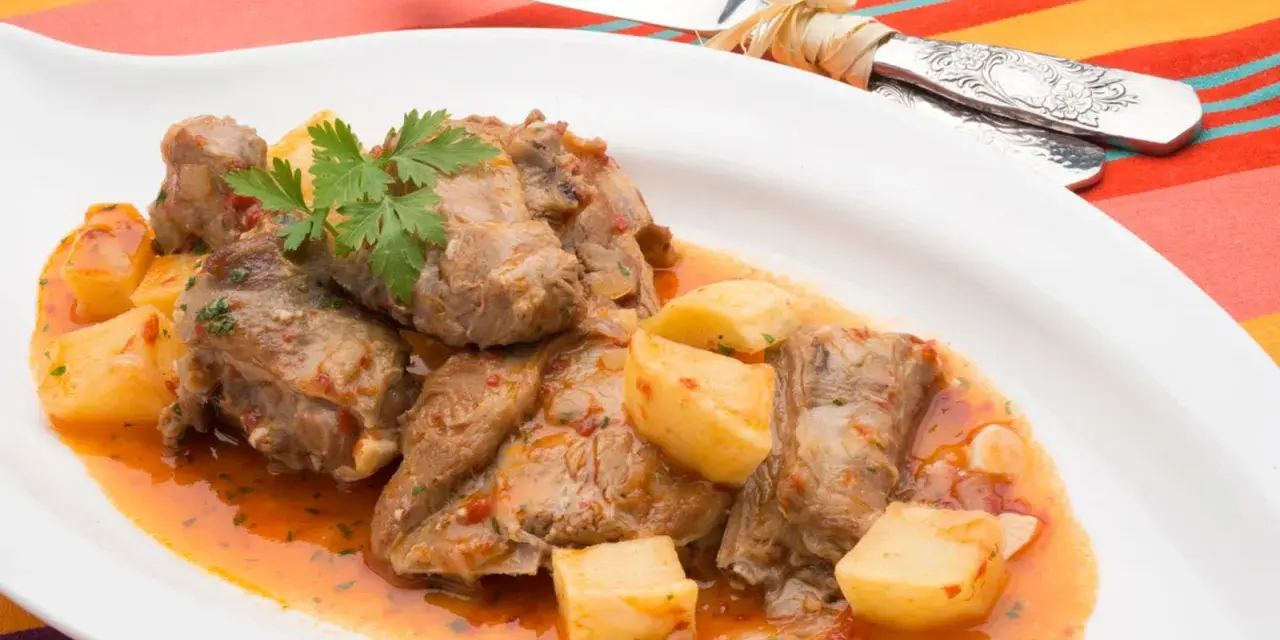 Sabroso cordero al chilindrón con patatas, servido en plato blanco con cubiertos atados con hilo.