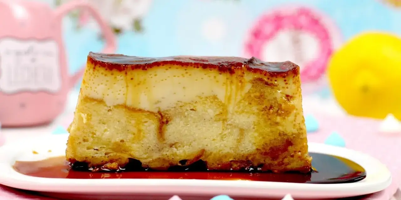 Un trozo de pan de calatrava, con su caramelo dorado, reposa en un plato blanco. Al fondo, una taza rosa y un limón.