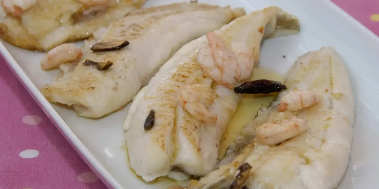 Filetes de gallo a la plancha con gambas y ajo laminado, servidos en una bandeja blanca.