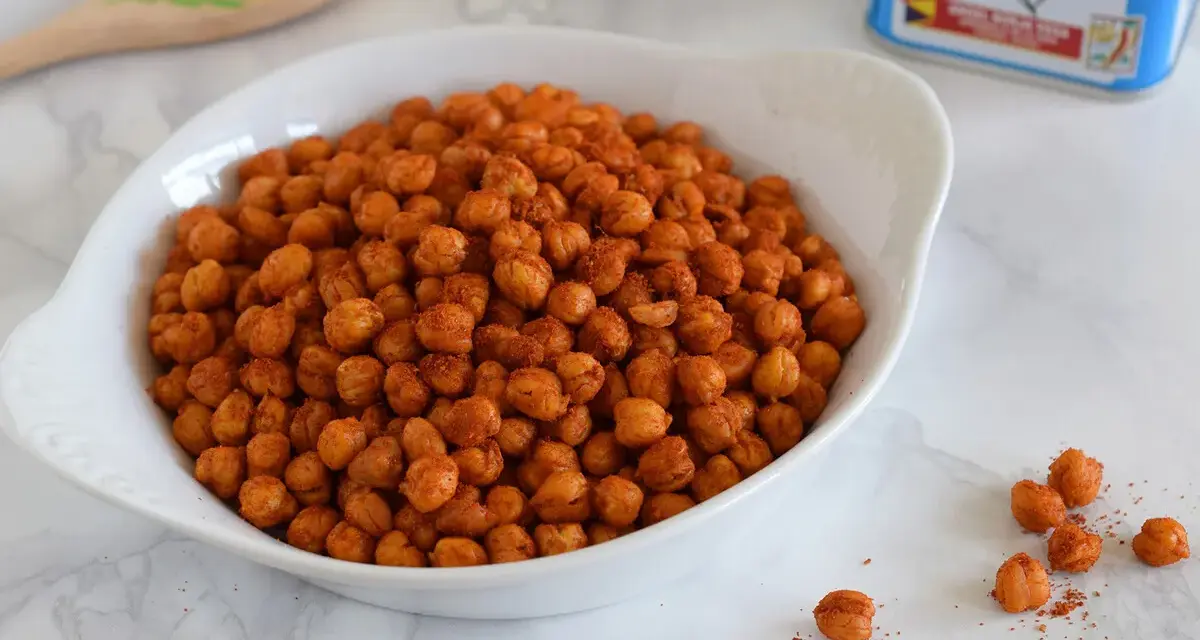 Un tazón blanco lleno de garbanzos fritos crujientes y especiados. Una lata de clavo y una cuchara de madera decorada están al fondo.