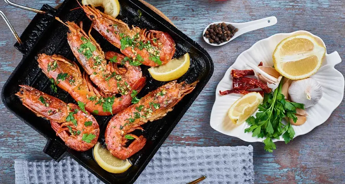 Deliciosos langostinos a la plancha, servidos con rodajas de limón y perejil fresco. Acompañados de ajo, guindillas y granos de pimienta.