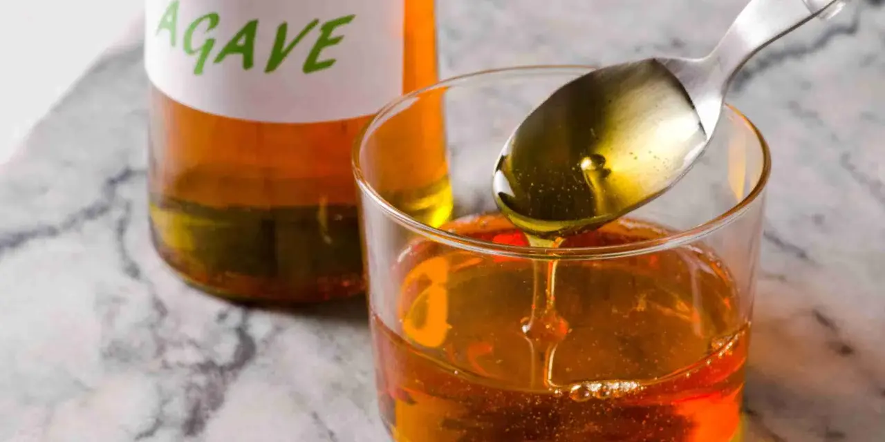 Una cuchara vierte sirope de agave dorado en un vaso lleno de este dulce líquido. Una botella con etiqueta "AGAVE" está al fondo.
