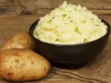 Un tazón de cremoso puré de patatas, adornado con perejil picado, junto a patatas enteras sobre una mesa de madera rústica.