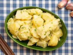Un tazón verde lleno de patatas alioli cremosas, adornadas con perejil fresco. A su lado, ajos y tenedores de madera sobre un mantel de cuadros azules.