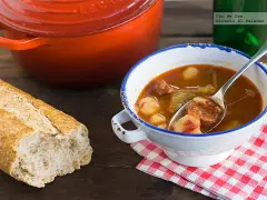 Un cuenco de patatas a la riojana humeante, con chorizo y pimientos, junto a una hogaza de pan rústico y una olla roja.