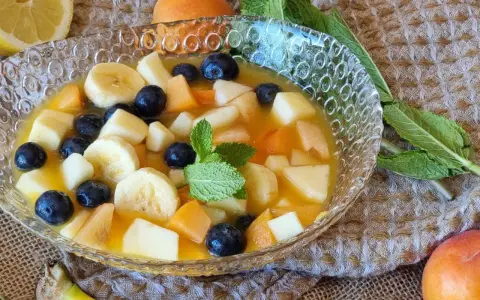 Deliciosa macedonia de frutas con plátano, arándanos y melocotón en un cuenco de cristal, decorada con menta.
