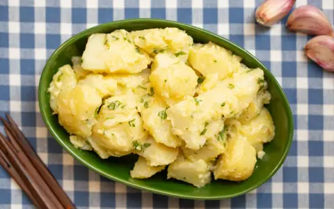 Un tazón verde lleno de patatas alioli cremosas, adornadas con perejil fresco. A su lado, ajos y tenedores de madera sobre un mantel de cuadros azules.