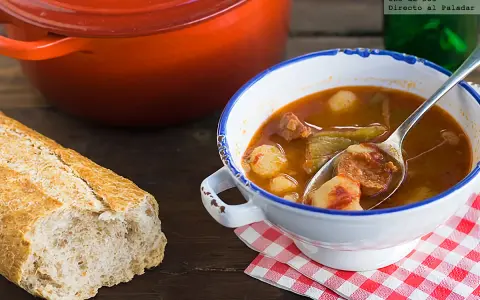 Un cuenco de patatas a la riojana humeante, con chorizo y pimientos, junto a una hogaza de pan rústico y una olla roja.