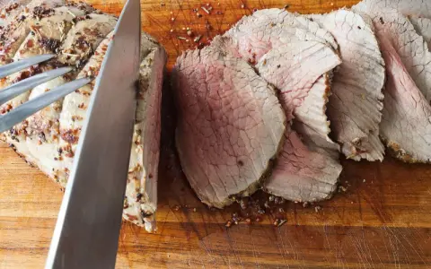 Un cuchillo corta un jugoso roast beef en rodajas sobre una tabla de madera.
