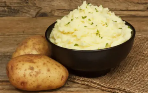 Un tazón de cremoso puré de patatas, adornado con perejil picado, junto a patatas enteras sobre una mesa de madera rústica.