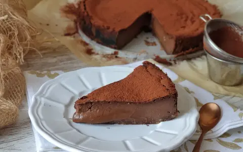 Una porción de tarta de chocolate, cubierta de cacao en polvo, reposa en un plato blanco. El resto de la tarta, también espolvoreada, se ve al fondo.