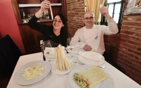 Pareja sonriente celebra con espárragos blancos en un restaurante. Platos de espárragos blancos y ensalada adornan la mesa.
