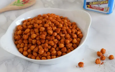 Un tazón blanco lleno de garbanzos fritos crujientes y especiados. Una lata de clavo y una cuchara de madera decorada están al fondo.