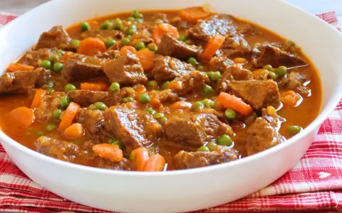 Ternera a la jardinera en un cuenco blanco, con trozos de carne tierna, zanahorias y guisantes en una salsa sabrosa.