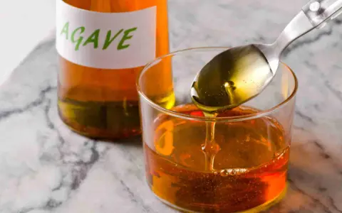 Una cuchara vierte sirope de agave dorado en un vaso lleno de este dulce líquido. Una botella con etiqueta "AGAVE" está al fondo.