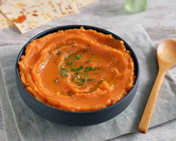 Un tazón de cremoso puré de boniato, adornado con aceite y hierbas, junto a una cuchara de madera.