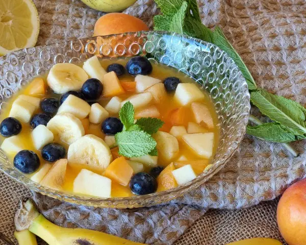 Deliciosa macedonia de frutas con plátano, arándanos y melocotón en un cuenco de cristal, decorada con menta.