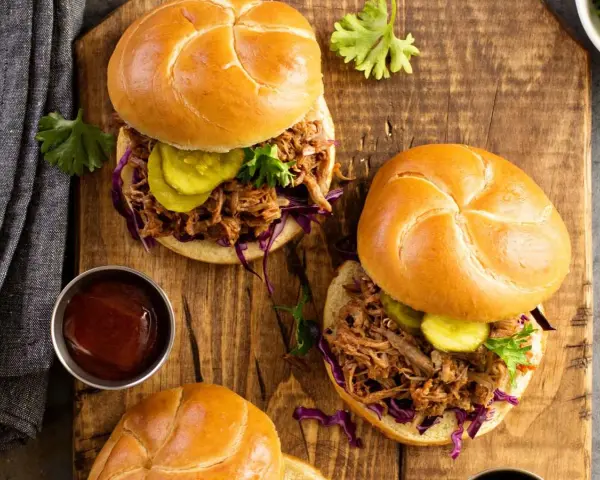 Dos sándwiches de pulled pork jugoso, con repollo morado, pepinillos y salsa barbacoa en un plato de madera.