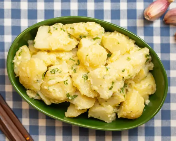 Un tazón verde lleno de patatas alioli cremosas, adornadas con perejil fresco. A su lado, ajos y tenedores de madera sobre un mantel de cuadros azules.