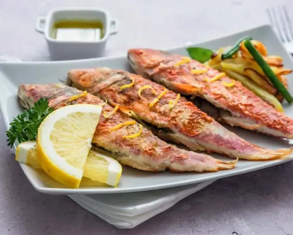 Cuatro salmonetes al horno, dorados y jugosos, servidos con rodajas de limón, perejil y patatas fritas.
