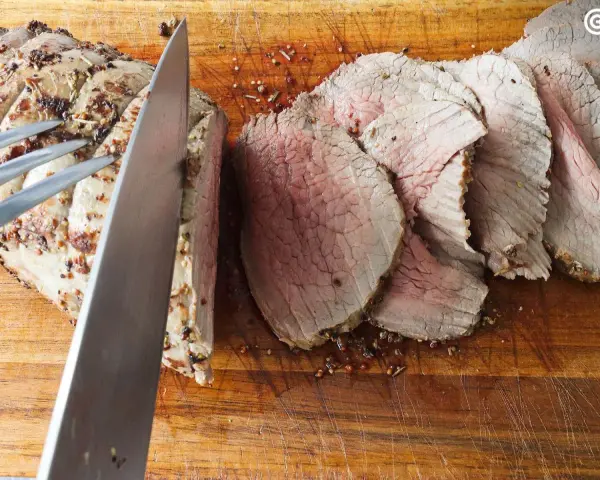 Un cuchillo corta un jugoso roast beef en rodajas sobre una tabla de madera.