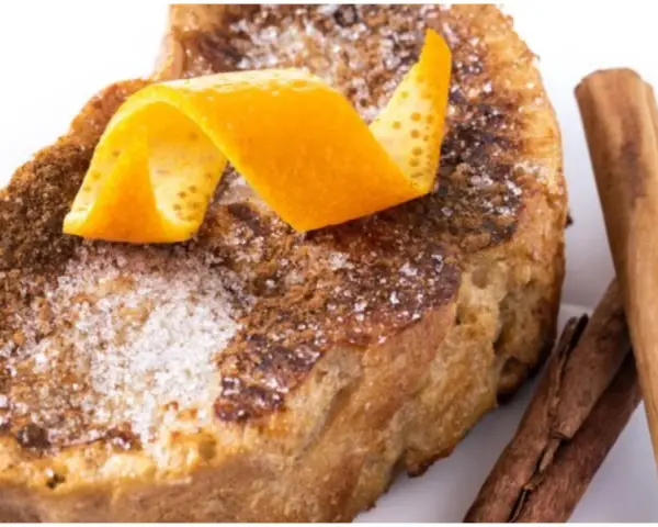 Torrijas al horno espolvoreadas con azúcar y canela, decoradas con piel de naranja y acompañadas de ramas de canela.