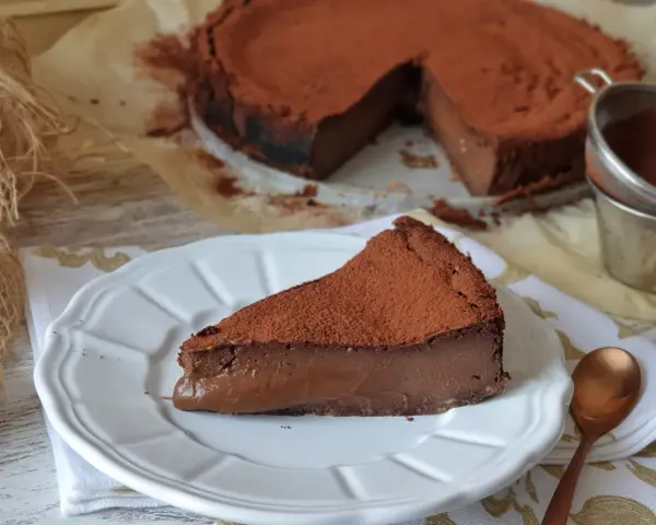 Una porción de tarta de chocolate, cubierta de cacao en polvo, reposa en un plato blanco. El resto de la tarta, también espolvoreada, se ve al fondo.